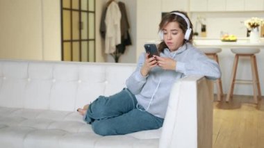 Modern bir dairede hafif bir koltukta oturan bir kız akıllı telefonuyla kulaklıklarıyla çalışıyor. Ciddi ve odaklanmış görünüyor. Arkasında modern bir mutfak ve cam kapılar var..