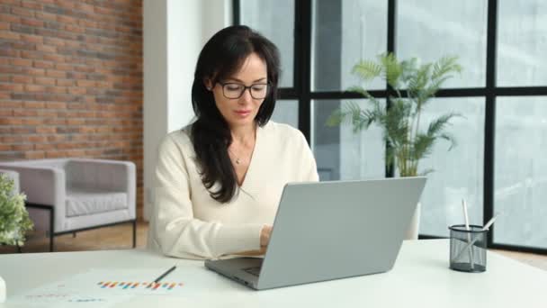 一个黑发女人在一间现代化的办公室的笔记本电脑上工作 她在电脑上比较纸质图表和电子图表 那个女人穿着眼镜和一件轻薄的毛衣 工作和企业 — 图库视频影像