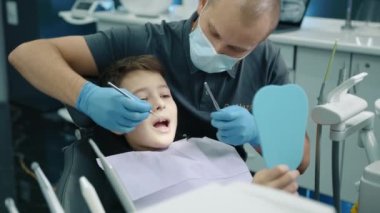 Sakin çocuk dişçi koltuğunda oturup ağzını kocaman açarken lateks eldivenler ve tıbbi maske uzmanı diş fırçalayıcı ve çukur aynasıyla dişleri kontrol ediyor. Yüksek kalite 4k görüntü