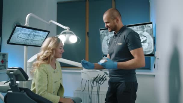 金发女士坐在牙医椅子上 看着牙科专家手拿着医用手套的牙科植入物样本 牙科治疗的现代设备 高质量的4K镜头 — 图库视频影像
