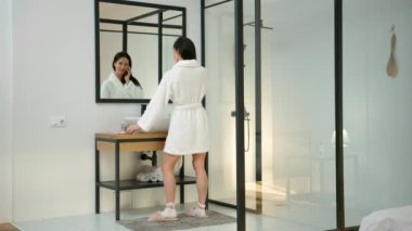 Beyaz cüppeli orta yaşlı bir kadın bir arkadaşıyla cep telefonunda konuşuyor. Aynanın önünde duruyor, saçını tarıyor, yürüyor ve sonra yatağa oturuyor. Minimalist stil.