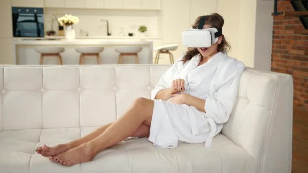 バーチャルリアリティグラスの幸せな女の子は 現代のテクノロジーの3D世界に没頭したモダンなアパートのソファーに座っています 彼女はVr体験で見たように 彼女の手でバーチャルオブジェクトを移動します — ストック動画