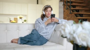 Genç bir kadın kulaklıkla müzik dinliyor ve akıllı telefon üzerinde çalışıyor. Modern bir dairede gri kapüşonlu bir koltukta oturuyor. Çiçekler ön planda kırmızı V-RAPTOR 8K RAW.