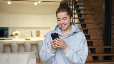 Gri kapüşonlu mutlu bir kız akıllı telefonuyla mesaj atıyor. İki katlı şık bir dairede oturuyor. Arka planda merdivenler, bir kanepe ve şık bir mutfak var. Kırmızı 8K RAW.