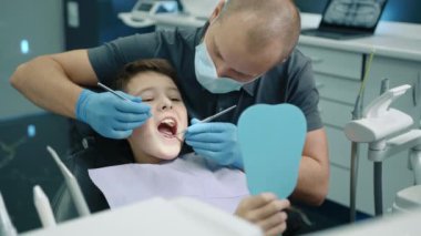 Dişçi koltuğunda oturan cesur çocuk, göğsünde peçeteyle, ağzı sonuna kadar açık, maske ve tıp eldivenlerinde diş fırçası ve çukur aynası kullanıyor. Yüksek kalite 4k görüntü
