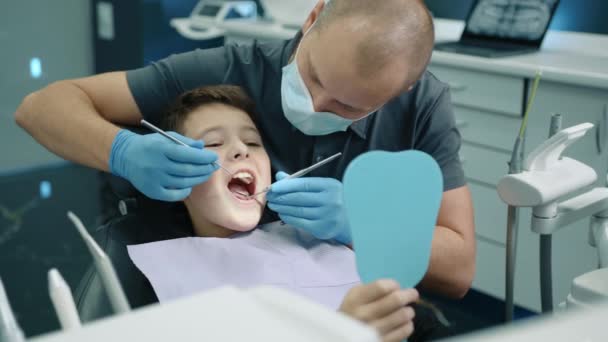胸にナプキン 口が広く開いた マスクと医療用手袋の歯科専門家が歯スクレーパー コンセーブミラーを使用して歯科チェアに座っている勇敢な少年 高品質の4K映像 — ストック動画
