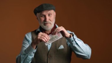 Yelekli ve siyah şapkalı bir emekli yüz dolarlık banknotu çarpıcı bir şekilde yırtıp atıyor. O, bileğinde klasik pahalı bir saatle mavi bir gömlek giyiyor. Kırmızı 8K RAW