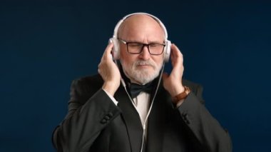 Smokinli şık yaşlı bir adam beyaz kulaklıklarla klasik müzik dinliyor. Yavaş hareket ediyor, müziğin tadını çıkarıyor. Gözlük takan sakallı adam. Koyu mavi arka plan. Kırmızı 8K RAW.