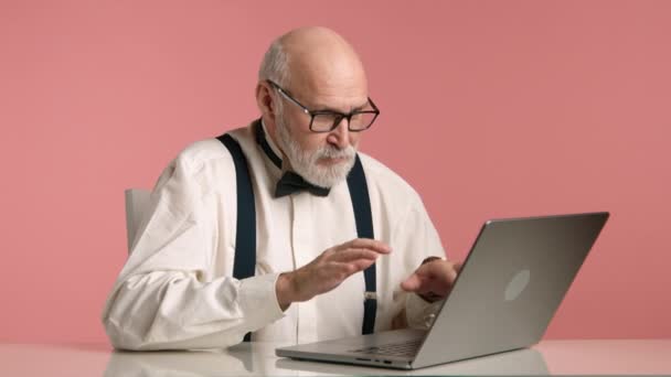 メガネと灰色のひげで60代のハゲな男がラップトップで作業を終え カメラで楽しそうに見える 彼はボウタイとサスペンサーで白いシャツを着ている ピンクの背景 Rawについて — ストック動画