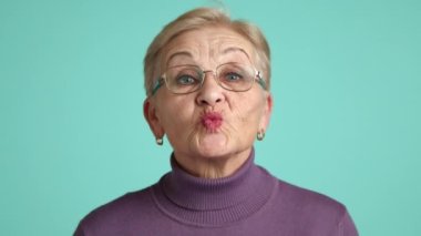 Kısa saç kesimi, derin kırışıklıkları, mor balıkçı yakalı hafif makyajı olan mutlu kadın emekliler stüdyoda mavi arka planda durup gülüyorlar. Yüksek kalite 4k görüntü