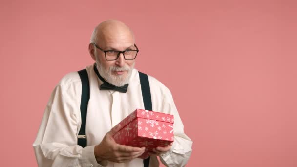 白いシャツにサスペンダーと弓のネクタイの男が赤いボックスにプレゼントをキャッチし それを開きます 彼の顔は 彼が見ているものから喜びをもって輝く ピンクの背景に対して サスペンサー付きのシャツを着た男 — ストック動画