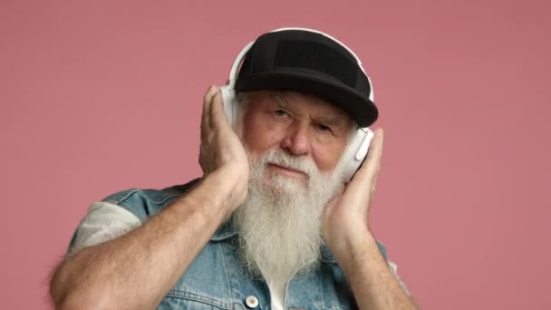 老年男子满头白胡子 高兴地微笑着 一边用白色耳机听着音乐 头戴黑色帽子 身穿牛仔背心 与粉色背景隔离 红色8K红色 — 图库视频影像
