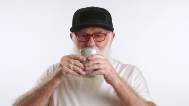 Nazik yaşlı adam, gri bardaktan çay içiyor. Siyah beysbol şapkalı ve son moda kırmızı gözlüklü büyükbaba. Beyaz bir arka planda beyaz bir tişört giymişti. Kırmızı 8K RAW.