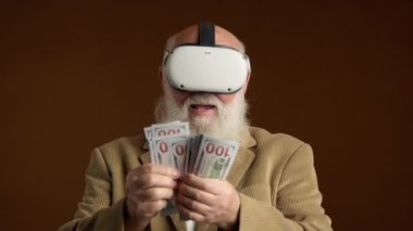 VR gözlüklü mutlu emekliler. Elindeki 100 dolarlık banknotları neşeyle sayıyor. O bir ceket giyer ve kahverengi bir arka plana karşı durur. 8K HAM.