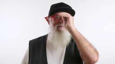 75 yaşını geçmiş yaşlı bir adam siyah beyzbol şapkasını, kırmızı gözlüğünü ve gülümsemesini çıkarıyor. Beyaz bir tişört ve siyah bir yelek giyiyor. Dedem beyaz arka planda duruyor. 8K HAM.