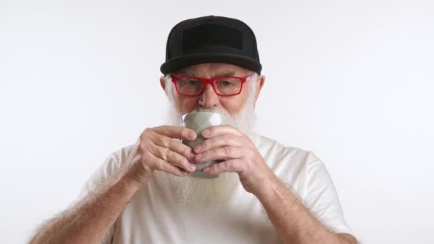 善良的老人 正在用灰色的杯子喝茶 祖父戴着黑色棒球帽 戴着时髦的红色眼镜 穿着白色T恤 背景是白色的 红色8K红色 — 图库视频影像