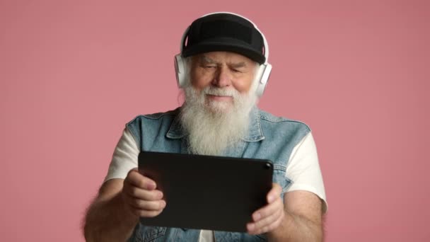 ハンサムな祖父はタブレットで積極的にゲームをしている 彼はティーンエイジャーのように振る舞い さまざまな感情を表現しています 白いヘッドフォン 黒い野球帽 大きな髭を着用しています デニム ベスト イギリス空軍 — ストック動画