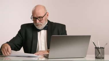 Gözlüklü ve sakallı ciddi bir iş adamı, bilgisayar üzerinde çalışıyor ve beyaz bir odada masa başında kağıtlarla çalışıyor. Haritaları inceliyor ve dikkatle inceliyor. Adam siyah takım elbise giymiş..