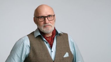 50 'li yaşlardaki seçkin bir adam kameraya ciddi ciddi bakıyor, sofistike bir insan. Onun klasik tüvit yelekli ve açık mavi gömleği açık gri arka planda göze çarpıyor. 8K HAM.