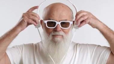 Beyaz güneş gözlüklü ve kulaklıklı, beyaz bir arka planda öne çıkan beyaz sakallı, yaşlı şık bir adam. Görünüşü zarafet ve zarafeti temsil ediyor. 8K HAM.