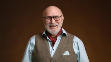 Orta yaşlı bir adam şık bir şekilde giyinmiş neşeli bir gülümsemeyle neşe ve mutluluk saçar. Gri bir sakalı var ve kahverengi bir arka planda duran klasik bir yelek ve boyun eşarbı giyiyor. Çiğ.