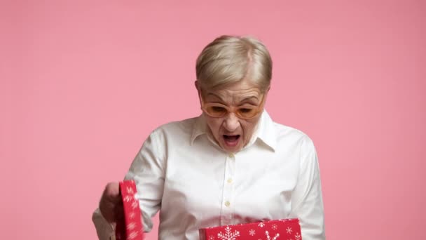 在打开用红纸包裹着雪花的盒子 站在粉色的背景上之后 这位穿着时髦眼镜白衬衫的老太太非常惊讶 高质量的4K镜头 — 图库视频影像
