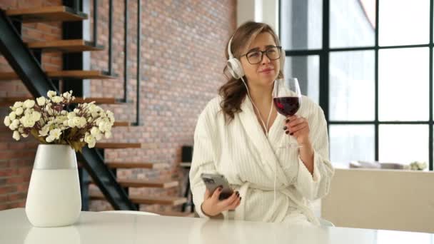 居心地の良い白いバスローブでリラックスした若い女性は 穏やかな瞬間を楽しんで 赤ワインを飲み スマートフォンをブラウジング モダンなロフトで 活気に満ちた花のディスプレイでアクセント — ストック動画