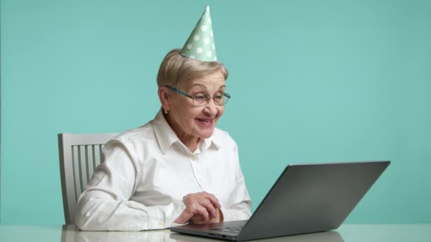 漂亮的老太太戴着眼镜 头戴生日帽 白衬衫 在笔记本电脑上有视频聊天 谈情说爱 坐在蓝色背景工作室之上 高质量的4K镜头 — 图库视频影像