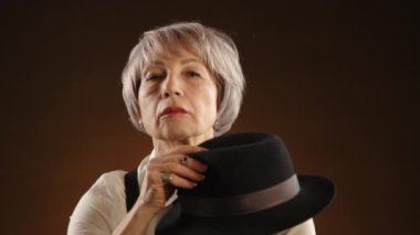Eski moda kıyafetler giymiş yaşlı bir kadın, pantolon askısı ve siyah kravat takan bir gömlek, siyah şapka. Şapkasını çıkardı ve kameraya baktı. Arka plan koyu kahverengidir. Kamera 8K HAM.