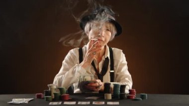 Yaşlı bir gangster kadın sigara içiyor ve pokerde kazandığı bir tomar parayı elinde tutuyor. O, pantolon askısı ve siyah şapkalı beyaz bir gömlek giydi. Kartlar ve fişler masada. 8K HAM.