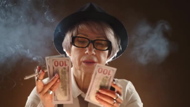 黒い帽子とメガネをかぶった高齢の女性は 優雅に片手に葉巻を持ち もう片方には100ドルの紙幣のファンがおり 彼女の神秘的な雰囲気が加えられています カメラ Raw — ストック動画