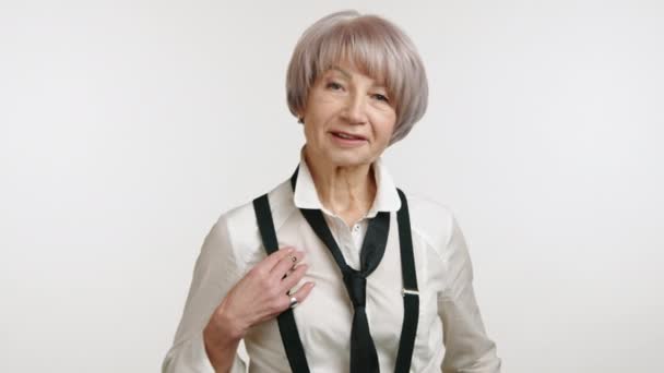 优雅的老年女性 有着时髦的银发 穿着时髦的白衬衫和经典的黑色吊带衫 自信地在中性的背景下摆出迷人的笑容 8K照相机 — 图库视频影像