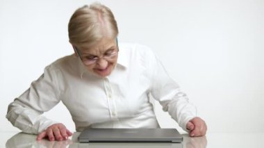 60-70 yaşlarında, gözlüklü, beyaz gömlekli, gülümseyen, laptopunu açan, ellerini ovuşturan, stüdyodaki dizüstü bilgisayarında yazı yazmaya hazırlanan sarışın bir kadın. Yüksek kalite 4k görüntü
