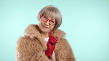 Modaya uygun yaşlı kadın kameraya neşe içinde poz veriyor. O, bej kürk ceket, kırmızı deri eldiven ve kırmızı gözlük takıyor. Kadın çok olumlu ve zarif. Yaşlılıkta stil ve güzellik. 8K HAM.
