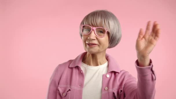 세련된 할머니는 따뜻하게 인사와 그녀는 분홍색 블라우스와 분홍색 배경에 안경에 — 비디오