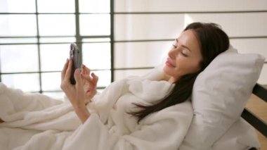 Yatak odasında yatağında mutlu bir kadın, telefonuyla sevinçle mesaj atıyor. Uzanır ve güzel beyaz bir bornoz giyer. Oda geniş pencerelerle dolu. Kamera 8K HAM.