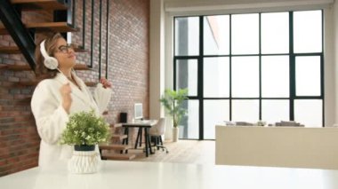  Evde yalnız kalmanın tadını çıkarıyorum. Bornozlu bir kız kulaklıkla müzik dinler ve büyük, güneşli pencereli, modern bir dairede dans eder. Kişisel alan. Kamera 8K HAM.