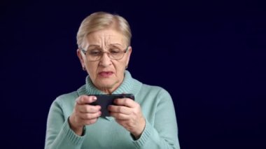 Kısa saçlı, mavi kazaklı, akıllı telefon ekranına tıklayarak video oyunu oynamaya takıntılı, kayıp ve zaferi ifade eden yaşlı sarışın kadın, stüdyo. Yüksek kalite 4k görüntü