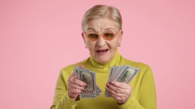 60-70 yaşlarında, bakımlı, iyi görünümlü, nakit, para sayan, ağzı açık, stüdyoda pembe arka planda duran yaşlı bir kadın. Yüksek kalite 4k görüntü