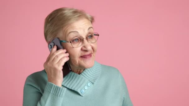 幸せな感情的なおばあちゃんは携帯電話で話します 青いセーターで短いブロンドの髪と陽気な女性年金 携帯電話を介して話す ピンクの背景 高品質の4K映像 — ストック動画