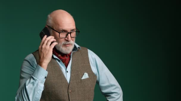 一位戴眼镜 留着胡子的老年绅士 在绿色的墙上愉快地通过电话交谈 他的服装典雅而精致 背心和领巾 8K照相机 — 图库视频影像