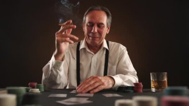 Poker oyununda kendine güvenen bir adam, ağzında bir puro ve elinde bir kart, poker fişi ve viski bardağına karşı hamlesini yapıyor. Yüksek kalite 4k görüntü