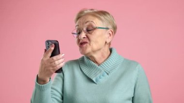 Neşeli, iyi görünümlü, bakımlı bir büyükanne cep telefonuyla video görüşmesi yapıyor, işaret parmağıyla işaret ediyor, tavsiye veriyor, el sallıyor, öpücükler yolluyor. Yüksek kalite 4k görüntü