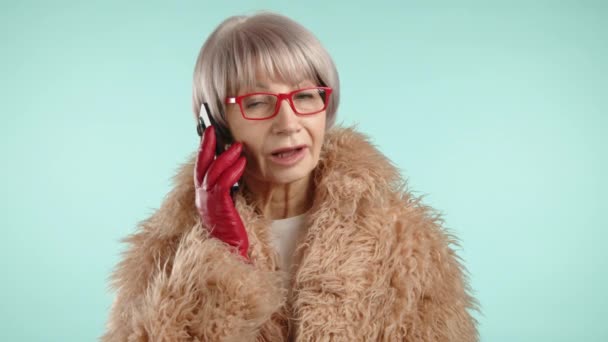 一位穿着毛皮大衣的老年妇女高兴地通过电话交谈 她有美丽的短发 红色的眼镜和红色的皮手套 为她的服装增添了一种特殊的风格 背景是蓝色的 8K照相机 — 图库视频影像