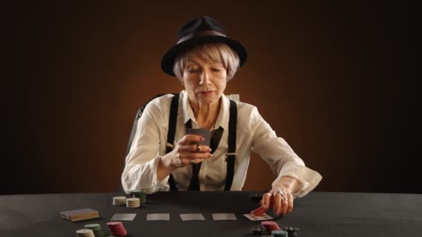 大人の女性が暗い部屋でポーカーをしている 彼女は勝利の組み合わせを見るためにテーブルの上にカードを持ち上げます ネクタイとサスペンサー付きの黒い帽子と白いシャツの女性 カメラ Raw — ストック動画