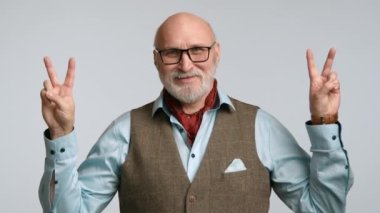 Şık bir tüvit yelek ve açık mavi gömlek içinde beyaz sakallı neşeli yaşlı bir adam, mavi bir arka planda yer alan geniş bir gülümsemeyle bir barış işareti veriyor. Kamera 8K HAM. 