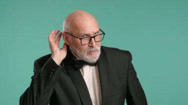 Keskin smokinli kıdemli bir adam, sanki işitme güçlüğü çekiyormuş gibi hareketler yapıyor, eli dikkatlice kulağının arkasına yerleştirilmiş, dingin bir donuk arka plana yaslanmış, bir merak hissi uyandırıyor. Kamera 8K HAM. 