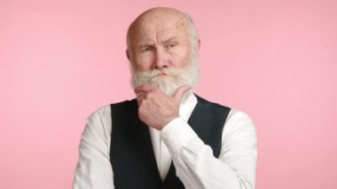 Çarpıcı beyaz sakallı ve düşünceli bir bakışla yaşlı bir adam çenesine dokunuyor, pastel pembe bir arka plana yaslanıyor, derin düşünce ya da karar verme mekanizması öneriyor. Kamera 8K HAM. 