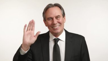 Terzi kıyafeti giymiş gülümseyen, olgun bir iş adamı, beyaz bir arka planda dostça bir jest yaparak sıcak bir şekilde selamlaşır. Kamera 8K HAM. 