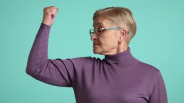 Neşeli, güzel, sarışın, kısa saçlı, gözlüklü, şaka yapan, güçlü bir adamın pozunda duran, mavi arka planlı, kazaklı, bisepsli bir kadın. Yüksek kalite 4k görüntü